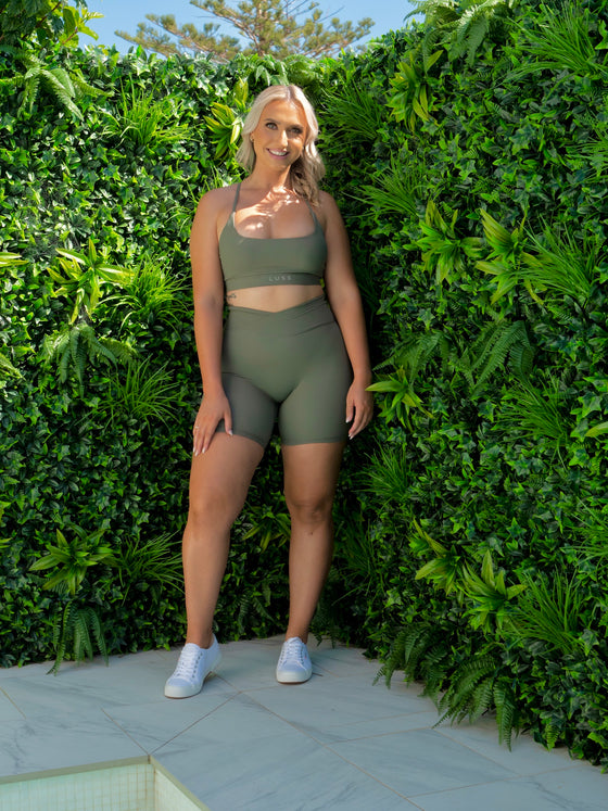 A photo of Maddi wearing the Sage Green Majestic bra styled criss cross at the back. Maddi is facing the camera and smiling. Shes infront of the green wall.