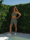 Mel from photoshoot day is semi turned to the side, with one hand on her waist. She is smiling and showing off her Sage Green Majestic Bra. She is standing infront of the green wall.