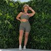 A photo of Makayla from shoot day in the Sage Green Majestic bra styled criss cross at the front. Shes touching her hair with one hand, and the other hand is on her waist facing the camera, infront of the green wall.