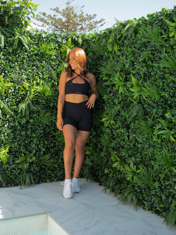 CHELSEA in the BLACK Lavish Bike Shorts and Majestic Bra styled criss cross at the front, head to the side smiling.