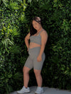 Yena in the Cloud Colour Bike Shorts (which is a light grey) and Majestic bra, styled criss cross at the back, infront of a green wall.